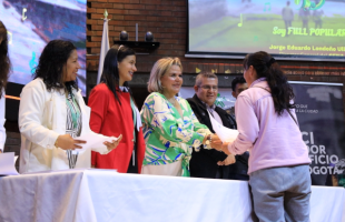 Varias personas detrás de una mesa hacen entrega de un diploma a una persona que está a espaldas de la cámara. 