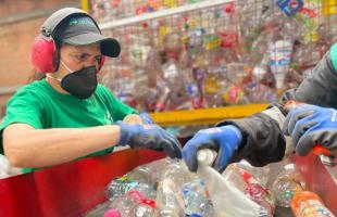 Foto en Interior - recicladora haciendo selección de material en una banda transportadora. La recicladora porta una camiseta verde, gorra negra con letra verde y cuenta con las medidas de protección para realizar esta labor. En el fondo se ve material plástico almacenado para hacer el tratamiento respectivo