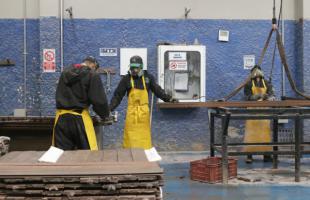 Tres personas con uniforme oscuro y delantal amarilla, trabajan en un taller de madera plástica.