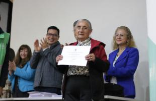  Foto  interior. Plano medio, se ve la mesa principal y hay personas que están entregando diplomas que en este momento están aplaudiendo a una mujer mayor, recicladora de oficio que está mostrando su diploma y posando para una foto.