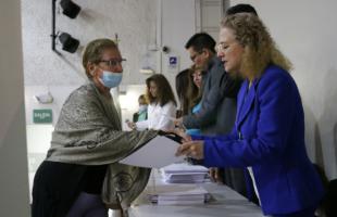 Foto  interior. Plano medio, se ve la mesa principal y hay personas que están entregando diplomas a los recicladores graduados. En primer plano sale una mujer con un vestido formal azul, cabello rubio y le está entregando a una mujer un diploma en sus manos.