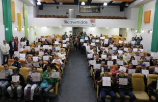  Foto al interior. Plano general de un auditorio en el que se ven personas sentadas a los dos lados levantando un diploma y al fondo se ven personas posando para la foto. Las paredes son blancas y cuenta con algunos detalles de colores verde, naraja, amarillo y verde.