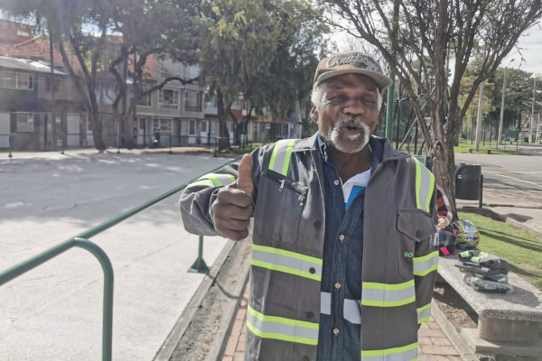 Foto exterior: se ve a un señor afrodescendiente en plano medio, porta un overol de color gris con reflectivos, una gorra negra y está en un parque. Tiene árboles en la parte trasera y al costado izquierdo de la foto se ve una foto